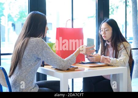 Junge kaukasische Frau zeigt ihrem asiatischen Freund nach dem Einkaufen etwas in ihrem Smartphone. Kaukasische und asiatische Frauen in einem Restaurant. Stockfoto