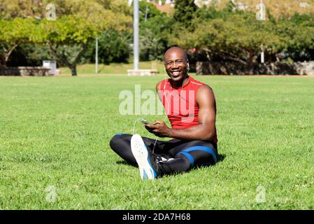 Porträt eines entspannten jungen afrikanischen Sportmanns, der im Freien sitzt Rasen und Musik hören Stockfoto