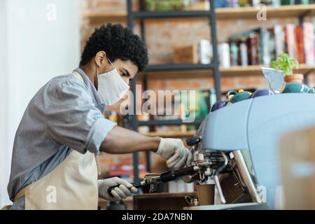 Beschäftigter junger afroamerikanischer Mann in Schutzmaske und weiß Handschuhe machen Cappuccino Stockfoto