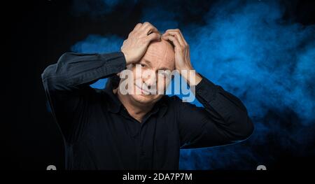 Alter Macho-Mann. Ein älterer Mann hält seine Hände hinter seinem Kopf auf einem dunklen Hintergrund mit blauem Rauch. Porträt eines älteren emotionalen Mannes auf einem dunklen Bac Stockfoto