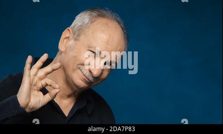 Alter Macho-Mann. Lächelnder älterer Mann zeigt mit seinen Händen eine gute Geste. Porträt eines älteren emotionalen Mannes auf dunklem Hintergrund im Studio Stockfoto