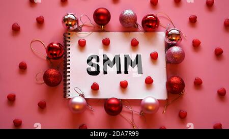 Die Inschrift SMM in einem Notizbuch auf einem rosa festlichen Hintergrund. Rote und rosa Weihnachtskugeln und Süßigkeiten in Form von Herzen Stockfoto