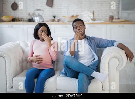 Nicotine abuse during pregnancy concept. Selfish black man smoking near his expectant wife at home Stock Photo