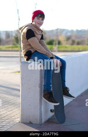 Kleiner Kapuzenjunge, der mit seinem Skateboard auf einem Zaun sitzt Stockfoto