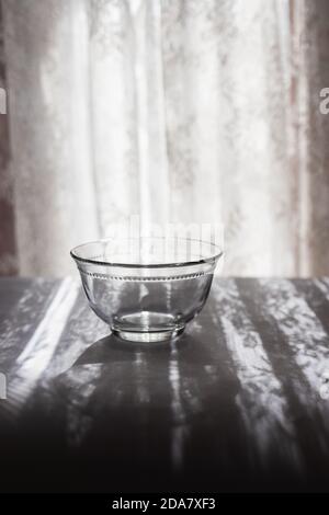 Leere Glasschale oder Tasse vor einem sonnigen Fenster mit einem Spitzenvorhängen und schönen Schatten auf dem Tisch. Einfache, minimalistische vertikale Aufnahme Stockfoto