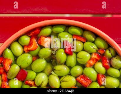 Schüssel frisch gepflückte grüne Oliven gewürzt mit rotem Pfeffer, Fenchel, Knoblauch und Thymian auf rotem Hintergrund Stockfoto