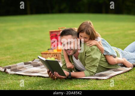 Neugieriges kleines Mädchen ablenkend, spielt mit ihrem Vater, wenn er ein Buch liest, während er auf einer Decke liegt. Glücklicher Vater und seine Tochter beim Picknick Stockfoto