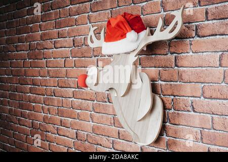 Intarsienhirsch an der Wand mit weihnachtsmannhut und roter Nase. Weihnachtsdekoration Stockfoto