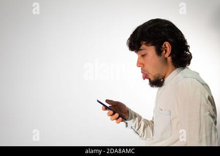 Junger hispanischer Mann in legerer Kleidung auf weißem Hintergrund Mit einem Handy und machen lustige Gesichter Stockfoto