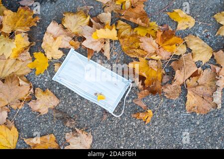 Gesichtsmaske auf einem Bürgersteig mit Herbstlaub in der Schweiz gedumpt. Die Maske wird verwendet, um die Coronavirus-Pandemie zu bekämpfen Stockfoto