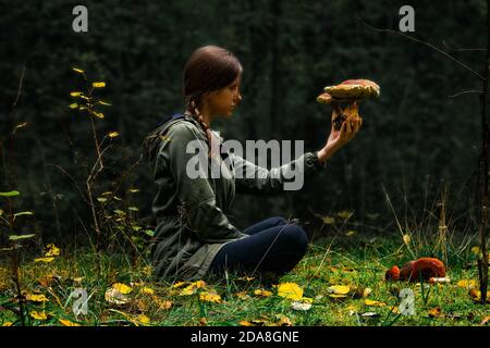 Eine Frau, die im Wald sitzt und die Wildpilze in der Hand hält und kontrolliert. Risiko einer Pilzvergiftung Konzept. Sichere Pilzjagd, Nahrungssuche, Pflücken. Stockfoto