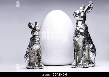 Zwei silberne Mate osterhasen Figuren vor einem weißen Keramikei, kreative ostern und Frühlingsdekoration 2020, Trendfarben auf weißem isoliertem Rücken Stockfoto