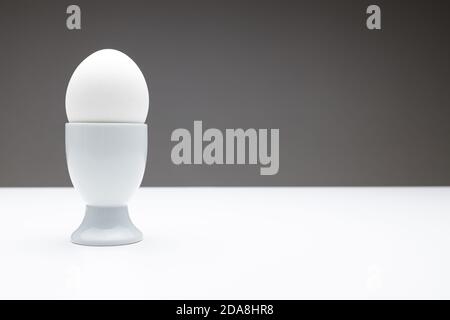 Vorderansicht eines Eierbechers mit einem weiß gekochten Ei auf einem dunkelgrauen Hintergrund auf einem weißen Tisch mit weichem Licht, als Vorlage für die Bemalung von Gesichtern oder Emoti Stockfoto