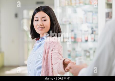 Fröhliche asiatische Frau, die Flasche mit Pillen in der Nähe Apotheker auf Verschwommener Vordergrund Stockfoto