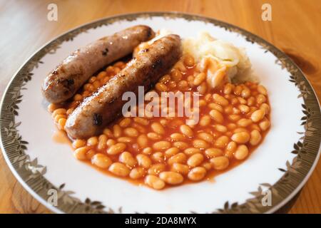 Zwei große Bratwürste mit gebackenen Bohnen und Kartoffelpüree auf einem weißen Teller mit goldenem Farbbesatz auf einem Holztisch, der von Tageslicht beleuchtet wird. Stockfoto