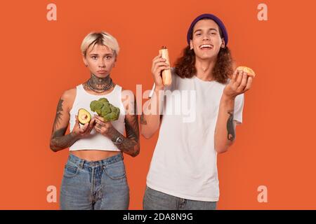 Freistil. Junge tätowierte Paar stehend isoliert auf orange Freundin hält Gemüse gesunden Lebensstil vs Freund hält Hot Dog und Donut Stockfoto