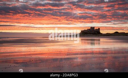 Ein lebhafter Sonnenaufgang spiegelt sich im nassen Sand am Bamburgh Beach wider, mit der silhouettierten Burg, die auf einem Felsvorsprung am Horizont thront. Stockfoto