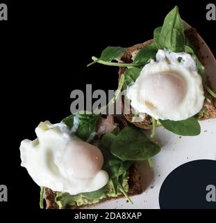 Pochierte Eier auf Toast mit Avocado, Speck und Spinatblättern, fotografiert vor schwarzem Hintergrund, mit Platz für Text. Stockfoto