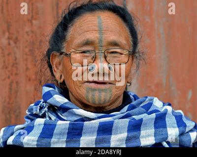 Alte nordöstliche indische Apatani Stammesfrau mit schwarzen hölzernen Nasenstopfen und traditionellem Gesichts-Tattoo trägt moderne Brillen und Posen für die Kamera. Stockfoto