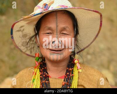 Ältere nordöstliche indische Apatani-Stammesfrau mit schwarzen Nasenstopfen und traditionellen Gesichts-Tattoos trägt einen modernen Sonnenhut und lächelt für die Kamera. Stockfoto