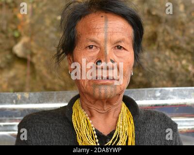 Ältere nordöstliche indische Apatani ethnische Minderheit Stammesfrau mit schwarzen hölzernen Nasenstopfen und traditionellen Gesichts-Tattoos Posen für die Kamera. Stockfoto