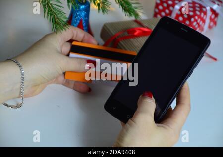 Frau kaufen Geschenke online mit Handy und Kreditkarte während der Weihnachtszeit. Handy mit Weihnachtsdekoration. Weihnachtsbaum und Stockfoto