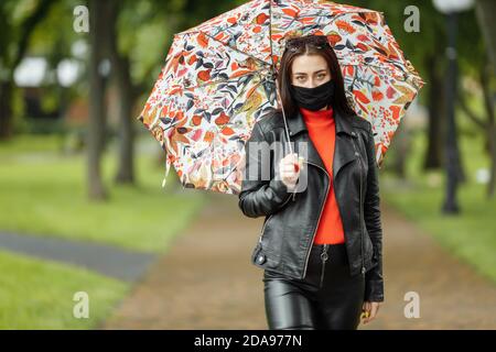 Ein maskiertes Mädchen läuft auf der Straße. Ein Mädchen in einer Schutzmaske geht im Park mit einem Regenschirm im Regen. Coronavirus-Infektion COVID-19 Stockfoto