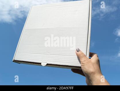 Eine Frauenhand hält eine weiße Pappschachtel mit einer Kopie des Raumes und des Platzes für Text auf einem blauen Himmel Hintergrund, Schablone, mockup Stockfoto