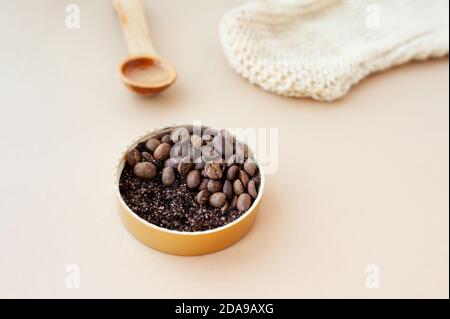 Kaffee Peeling große Granulat und Kaffeebohnen in Gold-Glas, kosmetisches Öl in Holzlöffel und raue Massage-Handschuh auf pastellbeige Hintergrund, Homema Stockfoto