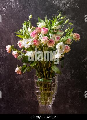 Eustoma und Rosen Bouquet in Vase auf texturiertem schwarzem Wandhintergrund. Stockfoto