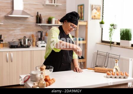 Ältere Frau trägt Schürze, während Mehl in Küchentisch bestreuen. Glücklicher älterer Koch mit einheitlicher Streuselung, sieben rohe Zutaten von Hand backen hausgemachte Pizza. Stockfoto