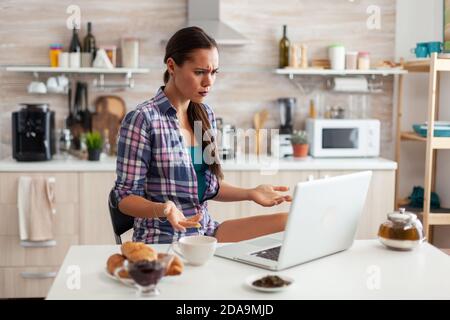 Stresed Frau mit Laptop in der Küche während des Frühstücks mit einer Tasse grünem Tee. Arbeiten von zu Hause mit Gerät mit Internet-Technologie, Surfen, Suchen auf Gadget am Morgen. Stockfoto