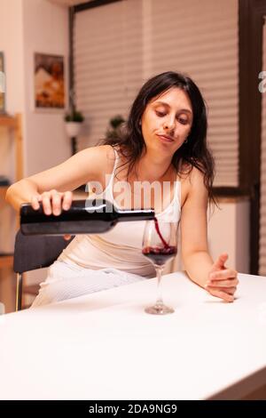 Unhapy Frau gießt Rotwein aus Flasche in Glas allein sitzen am Tisch in der Küche. Unglückliche Person Krankheit und Angst Gefühl erschöpft mit Alkoholismus Probleme. Stockfoto