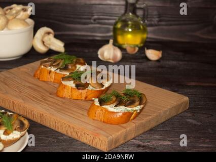 Baguette Toast mit weichem Frischkäse und Pilzen auf einem Holzbrett. Öl, Knoblauch und Pilze im Hintergrund. Snacks, schnelles Frühstück. Stockfoto