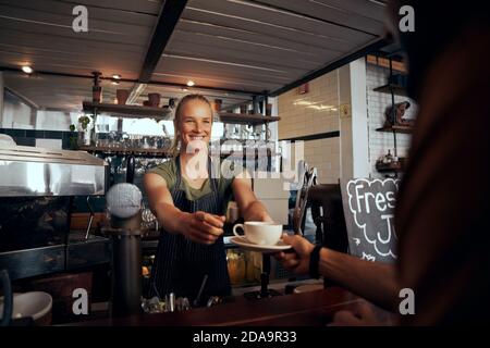 Schöne junge Kellnerin, die dem Gast eine Tasse Kaffee serviert In einem modernen Café Stockfoto