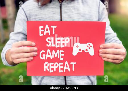 Männliche Teenager Gamer hält ein Banner mit essen, schlafen, Spiel, wiederholen Zitat Nachricht mit einem Gamepad-Symbol. Spielabhängigkeit oder Abhängigkeitskonzept. Stockfoto