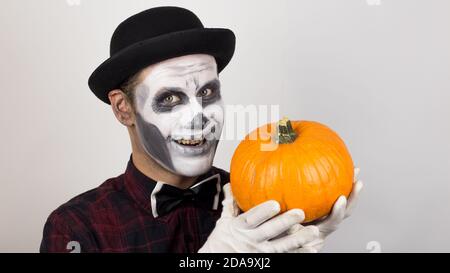 Ein gruseliger Clown schaut auf die Kamera, hält einen Kürbis in seine Hände und bedroht sie mit einem Messer. Halloween Kostüm. Stockfoto