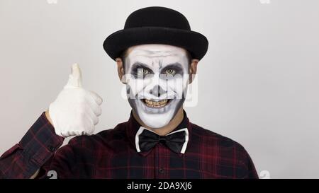 Ein schrecklicher Mann in Clown Make-up lächelt und zeigt einen Daumen nach oben. Ein beängstigender Clown schaut auf die Kamera und lacht schrecklich. Stockfoto