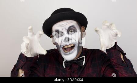 Ein schrecklicher Mann im Clown Make-up grimaces und macht erschreckende Gesten. Ein beängstigender Clown schaut auf die Kamera und lacht schrecklich. Stockfoto