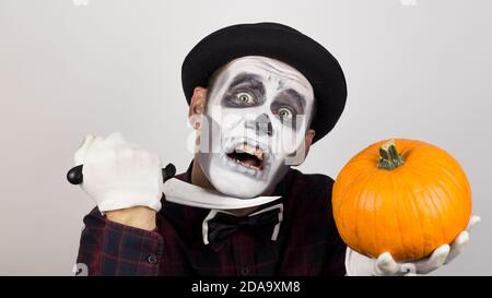 Ein gruseliger Clown schaut auf die Kamera, hält einen Kürbis in seine Hände und bedroht sie mit einem Messer. Halloween Kostüm. Stockfoto