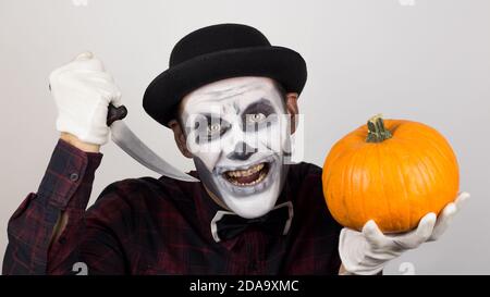 Ein gruseliger Clown schaut auf die Kamera, hält einen Kürbis in seine Hände und bedroht sie mit einem Messer. Halloween Kostüm. Stockfoto