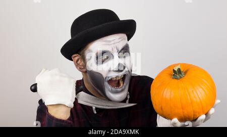 Ein gruseliger Clown schaut auf die Kamera, hält einen Kürbis in seine Hände und bedroht sie mit einem Messer. Halloween Kostüm. Stockfoto