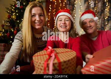 Eine glückliche Familie mit Kind in roten Weihnachtsmütze feiert Weihnachten. Nettes Mädchen hält verpacktes Geschenk, lächelnd und winkende Hand Stockfoto