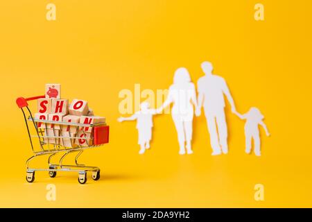 Wagen gefüllt mit Holzwürfeln, mit der Inschrift Shopping. Gelber Hintergrund mit einer Familiensilhouette aus Papier geschnitten. Speicherplatz kopieren. Das Konzept von Stockfoto