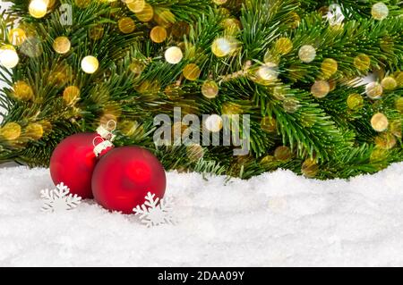 Nahaufnahme von zwei roten Weihnachtskugeln und Schneeflocken auf Schnee mit Tannenzweigen und Bokeh-Lichtern im Hintergrund. Weihnachtsschmuck als Gruß Stockfoto