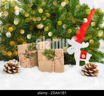 Zwei handgefertigte Weihnachtsgeschenke ohne Plastik, Holzrentiere im Weihnachtsmütze und Zapfen im Schnee mit Kiefernästen und Bokeh-Lichtern als Hintergrund. W Stockfoto