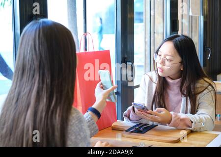 Junge kaukasische Frau zeigt ihrem asiatischen Freund nach dem Einkaufen etwas in ihrem Smartphone. Kaukasische und asiatische Frauen in einem Restaurant. Stockfoto