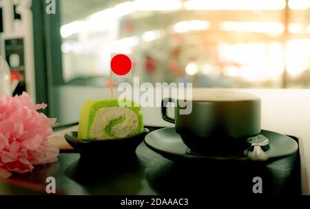 Heiße Kaffeetasse mit Scheibe grüner schweizer Rollkuchen mit Sahne auf dem Teller im Café auf Holzplatte im Café. Latte, Cappuccino oder Espresso in Stockfoto