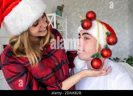 Junge verspielte Frau dummend um die Dekoration Mann mit Weihnachtskugeln statt Weihnachtsbaum. Paar tragen weihnachtsmützen und roten Schlafanzug. Stockfoto