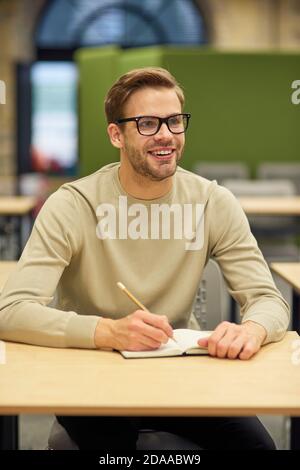 Business Education Konzept. Fröhlicher männlicher Büroangestellter sitzt am Schreibtisch im Coworking Space, macht Notizen und lächelt während des Corporate Trainings oder Seminars Stockfoto